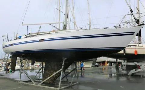Un grand voilier blanc et bleu est sur des chandelles, son flanc couvert de bernacles. Un homme nettoie la coque d'un autre bateau à proximité.