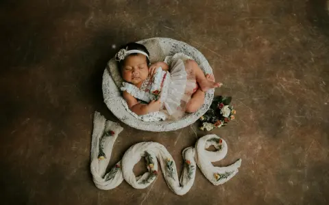 Dolce foto neonato 1 mese dorme in una cesta, circondato da fiori e dalla parola "love" creata con tessuto.
