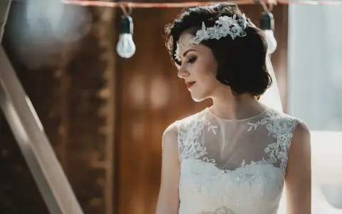 Panna młoda z krótkimi włosami w stylu boho, ozdobionymi kwiatową opaską, w białej sukni z koronkowymi detalami.