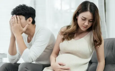 Ein Mann mit verdecktem Gesicht sitzt abgewandt von einer schwangeren Frau. Die Szene deutet auf eine Trennung in der Schwangerschaft hin, die mit Kummer verbunden ist.