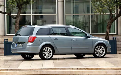 Srebrny Opel Astra H kombi zaparkowany przed nowoczesnym budynkiem z drzewami.