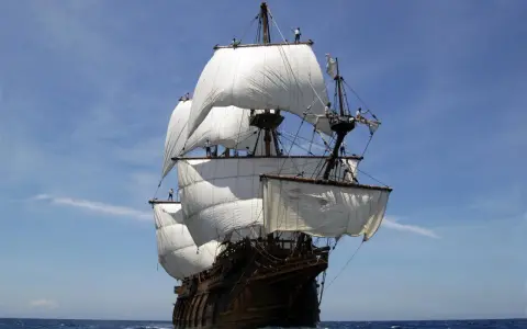 Un majestueux galion espagnol navigue sous un ciel bleu, ses voiles blanches gonflées par le vent.
