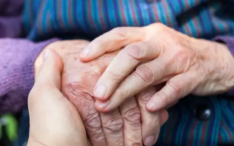 Dłonie młodszej osoby obejmują dłoń starszej, symbolizując wsparcie w obliczu choroby Alzheimera i jej przyczyn.