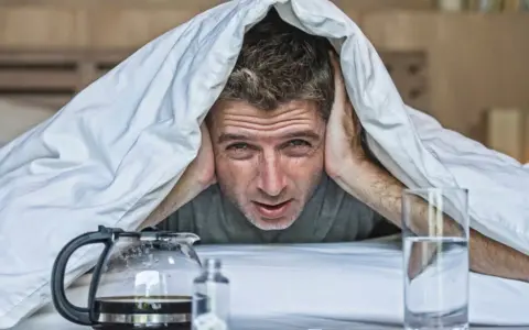Mężczyzna pod kołdrą z rękami na uszach, z kawą i wodą obok. Wygląda na to, że ma uczucie kaca bez alkoholu.