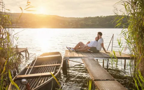 Para, gdzie na Mazury we dwoje? Para siedzi na pomoście, łódka przy brzegu, zachód słońca.