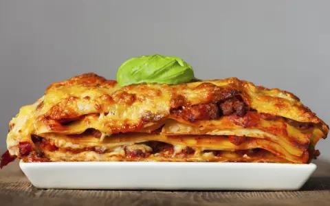 Una porción generosa de lasaña de carne, con capas de pasta, salsa y queso dorado, coronada con una hoja de albahaca fresca.