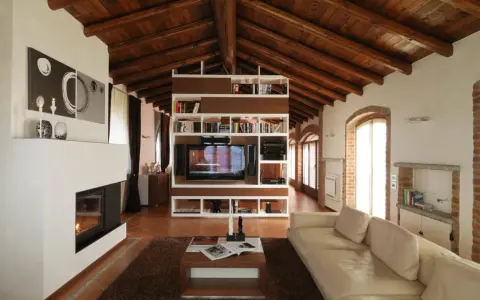 Accogliente soggiorno mansardato con soffitto in legno a vista, camino moderno e libreria centrale. Idee mansarde in legno per un ambiente caldo.
