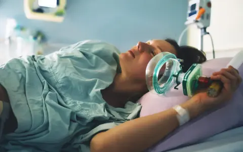 Kobieta w szpitalnym łóżku, z maską gazu rozweselającego przy twarzy, przygotowuje się do porodu.