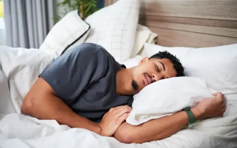 Ein Mann schläft tief und fest, um sein HRV zu verbessern. Er liegt auf einem weißen Bett mit vielen Kissen.