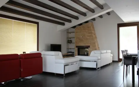 Soffitto con travi in legno scuro, un camino in pietra e divani bianchi. Ecco come fissare travi in legno al soffitto per un effetto rustico.