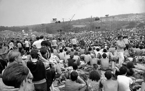 Tysiące ludzi siedzi na trawie, patrząc na scenę festiwalu. To jest Woodstock, gdzie jest muzyka i wolność.