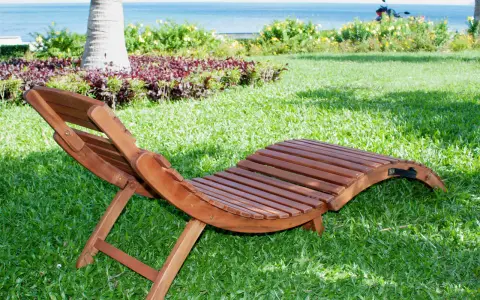 Chaise longue pieghevole in legno su prato verde, con mare e palme sullo sfondo. Ideale per rilassarsi all'aperto.