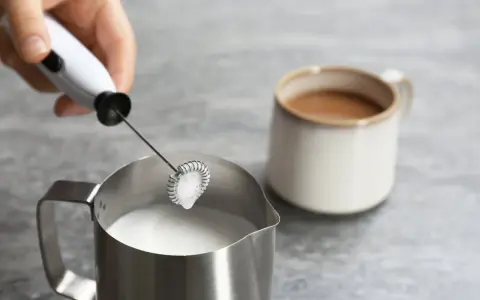 Hand hält Milchaufschäumer über Metallkännchen mit Milch. Perfekt für leckere Milchschaum Rezepte.