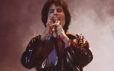 Freddie Mercury śpiewa z pasją, czując, że chce się uwolnić. Błyszcząca kurtka odbija światła sceny.
