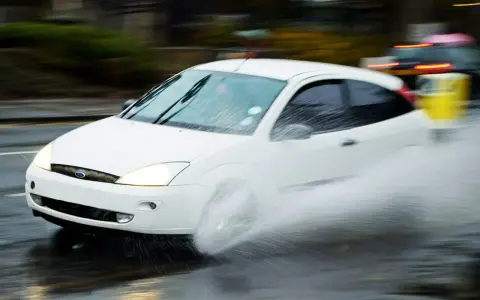 Auto bianco in movimento su strada bagnata, con un'enorme spruzzata d'acqua che indica l'aquaplaning cos'è.