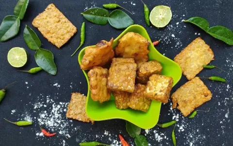 Knusprig gebratener Tempeh, ein fermentiertes Sojaprodukt, liegt in einer grünen Schale. Dazu garnieren Limettenscheiben, grüne Chilis und Blätter.