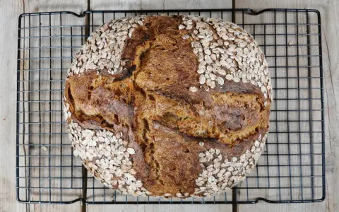 Ein rustikales Porridge Brot mit Haferflockenkruste, das auf einem schwarzen Gitter auf Holzdielen abkühlt.