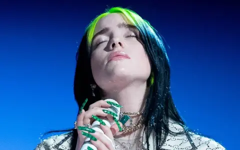 Piosenkarka Billie Eilish z zamkniętymi oczami śpiewa do mikrofonu, jej zielone pasemka włosów lśnią.