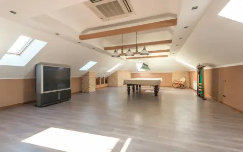 Vaste pièce sous les combles aménageables, avec table de billard, TV, fauteuil à bascule et étagères. Idéal pour un espace de loisirs.