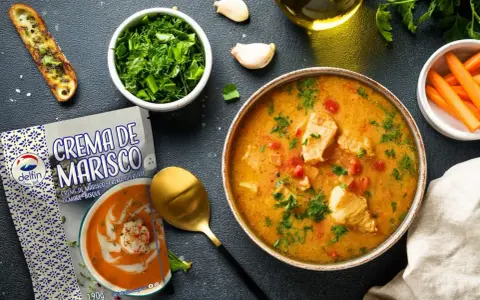 Un tazón de sopa de pescado casera con trozos de pescado, tomates y perejil, junto a una tostada, zanahorias y un paquete de crema de marisco.