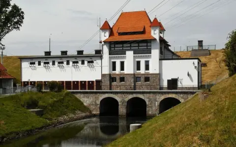 Elektrownia wodna Straszyn z charakterystycznym czerwonym dachem i kamiennymi arkadami nad kanałem.