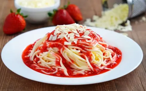 Ein Teller mit Spaghetti Eis, garniert mit Erdbeersauce und weißen Schokostückchen. Frische Erdbeeren und geriebene Schokolade im Hintergrund.