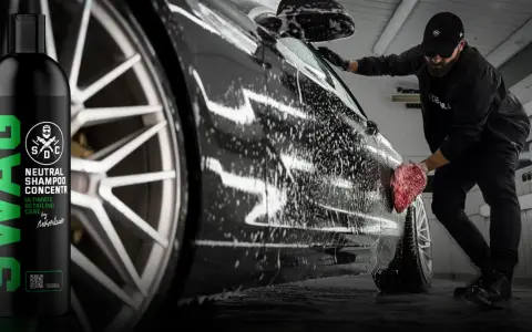 Mężczyzna myje czarne auto pianą, używając gąbki. Obok butelka szamponu SWAG.
