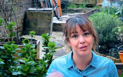 Mujer sonriente en un jardín con plantas verdes y macetas.