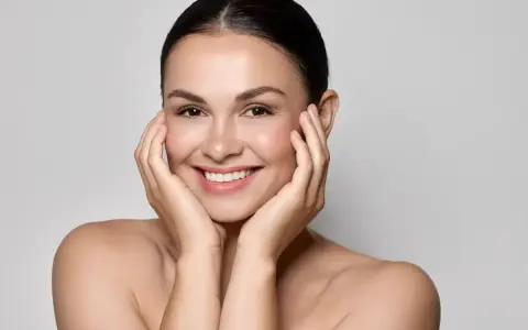 Uśmiechnięta kobieta z idealną, lśniącą cerą typu "glass skin", delikatnie dotykająca policzków dłońmi.