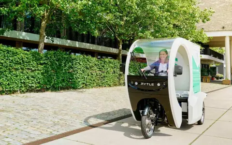 Eine Frau fährt ein Rytle Movr3, ein dreirädriges Elektrofahrzeug, vorbei an einer grünen Hecke und einem Gebäude.