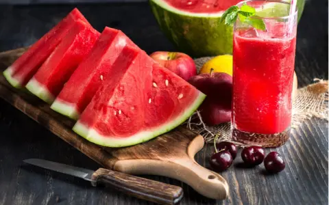 Saftige Wassermelone-Scheiben und ein Glas erfrischender Wassermelonensaft, reich an Vitaminen. Äpfel und Kirschen ergänzen das gesunde Arrangement.