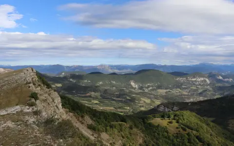 Vue panoramique depuis le col de Perty, avec des montagnes verdoyantes et un ciel bleu parsemé de nuages.