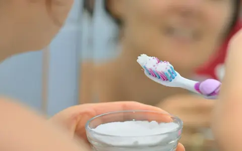 Ręka trzymająca szczoteczkę do zębów z białym proszkiem, obok miseczka z sodą oczyszczoną. Domowa pasta do zębów gotowa.