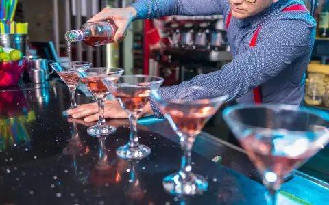Barman nalewa różowy drink do kieliszków koktajlowych na czarnym blacie.