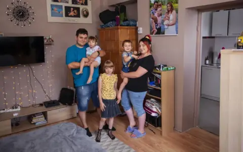 Rodzina z trójką dzieci pozuje do zdjęcia w salonie. W tle widać zdjęcia, telewizor i ozdobne lampki. Ich życie na kredyt nie przeszkadza im w cieszeniu się chwilą.