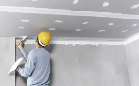 Pracownik w żółtym kasku nakłada masę szpachlową na sufit, przygotowując go do montażu. To kluczowy etap, by wiedzieć, jak przykleić styropian do sufitu.