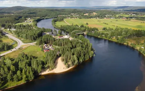 En vacker vy över en svensk landsbygd med en flod som slingrar sig genom gröna fält och skogar. En bro spänner över vattnet, och små röda hus syns bland träden. En idyllisk bild av sommar i Sverige.