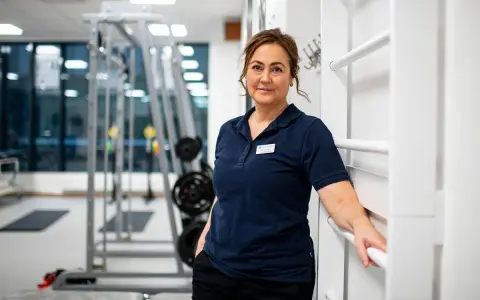 En kvinna i träningskläder står i ett gym. Hon har en namnskylt på sin tröja. I bakgrunden syns träningsredskap, som kan användas för att stärka underbenets muskler.
