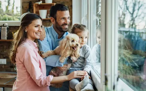 Szczęśliwa rodzina zastępcza, z pieskiem w ramionach, patrzy przez okno. Marzą o adopcji i wspólnym życiu.