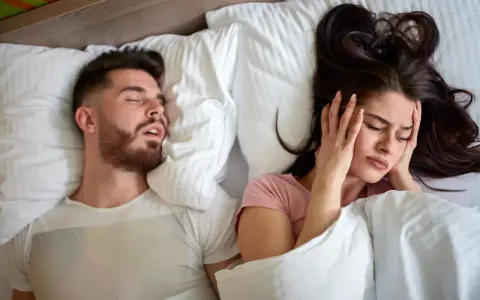 Kobieta zatyka uszy, próbując zasnąć obok męża, który głośno chrapie. Może czas na wizytę u lekarza od chrapania?