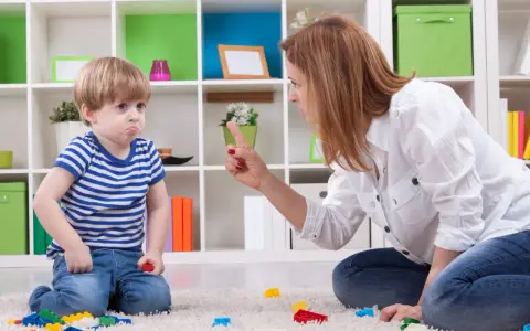 Mama karci synka za kłamstwo. Chłopiec ma smutną minę, a na dywanie leżą klocki.