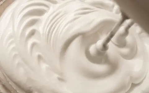 Ein Handmixer schlägt gerade cremige Sahne. Die weiße Masse bildet sanfte Wellen und Spitzen.