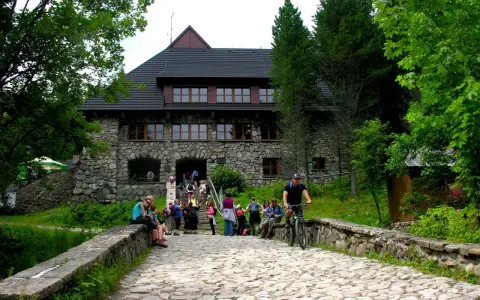 Kamienny budynek schroniska w Dolinie Chochołowskiej z ludźmi na schodach i rowerzystą na kamiennej drodze.