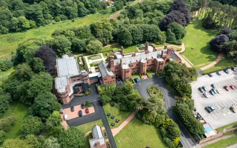 Aerial view of the abbey guest house, a grand brick building surrounded by lush green trees and manicured lawns. Cars are parked in a lot.