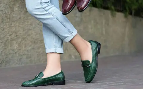 Un mocassino verde con abito elegante, perfetto per un look sofisticato. Un altro paio marrone è tenuto in mano.