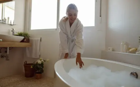 Kąpiel w rumianku na co pomaga? Kobieta w szlafroku dotyka piany w wannie, przygotowując relaksującą kąpiel.