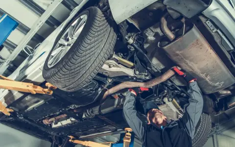 Mechanik pracuje pod samochodem, naprawiając budowę układu kierowniczego. Widać podniesione auto, koło i elementy wydechu.