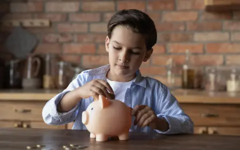 Niño introduce una moneda en una hucha rosa, aprendiendo sobre el ahorro con la cuenta infantil BBVA.