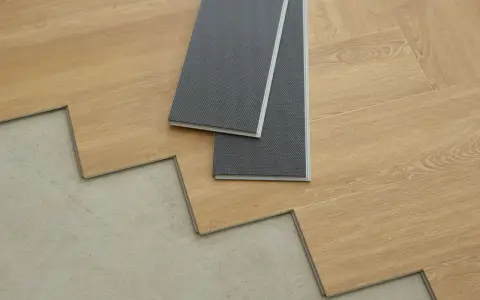 Panele winylowe w trakcie układania na betonowej podłodze. Dwa szare panele leżą na drewnianych deskach, pokazując, jak układać panele winylowe.