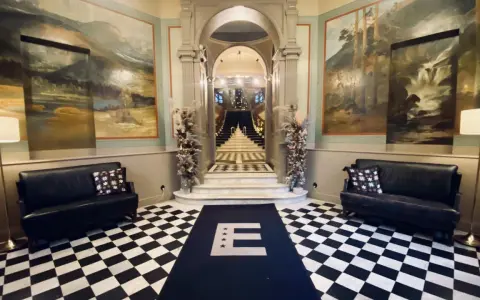 Elegant foajé med schackrutigt golv, konstverk på väggarna och en trappa. En matta med bokstaven E leder mot trappan. Känns som en plats med mycket hotell Knaust historia.